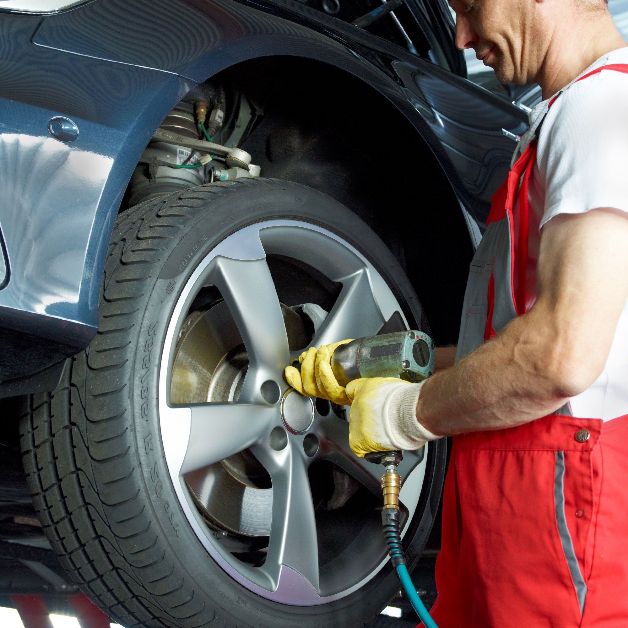 Mann in roter Latzhose arbeitet an einem Auto, Reifen wird mit Werkzeug bearbeitet.
