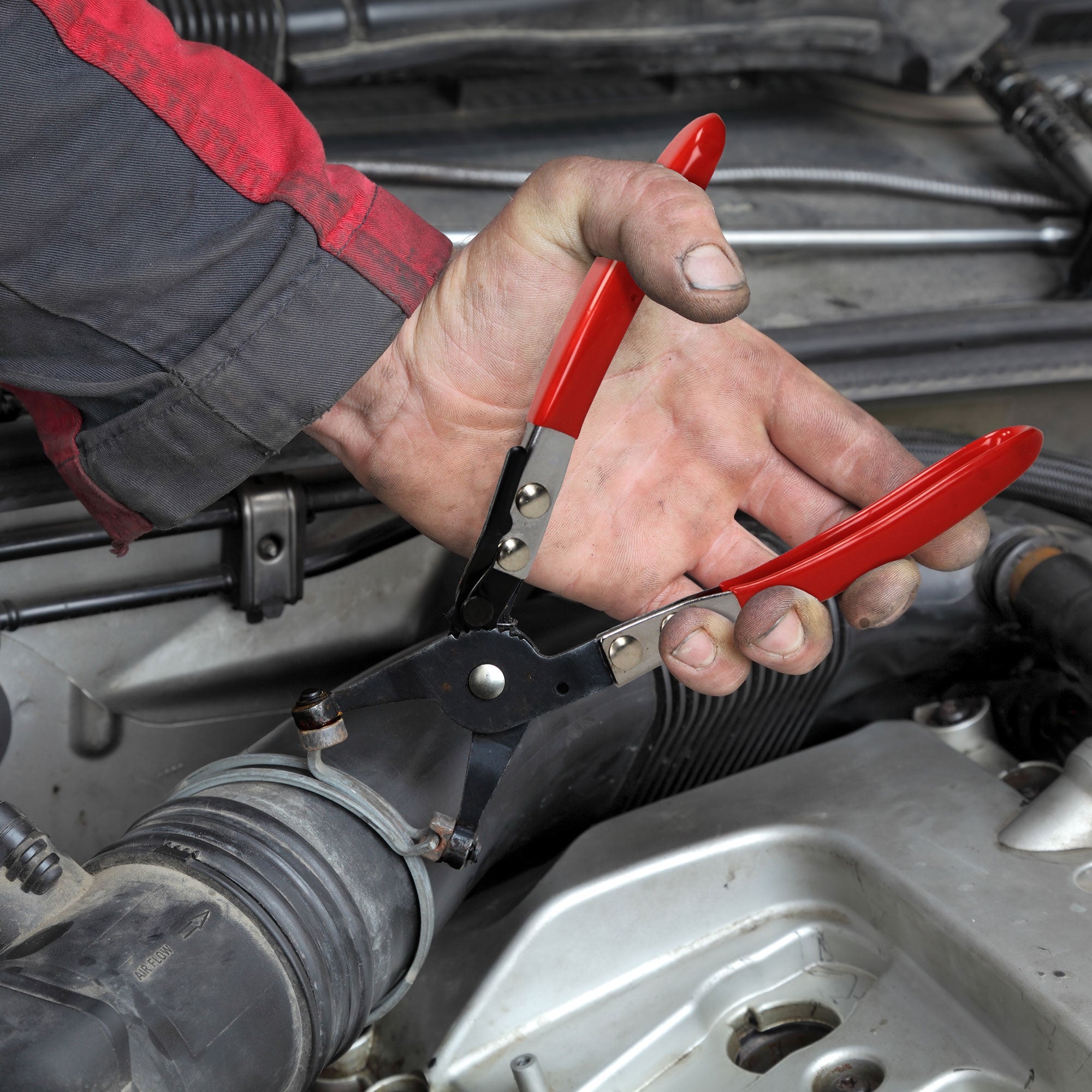 Hand mit Zange über einem Motorraum, rote und schwarze Arbeitskleidung.