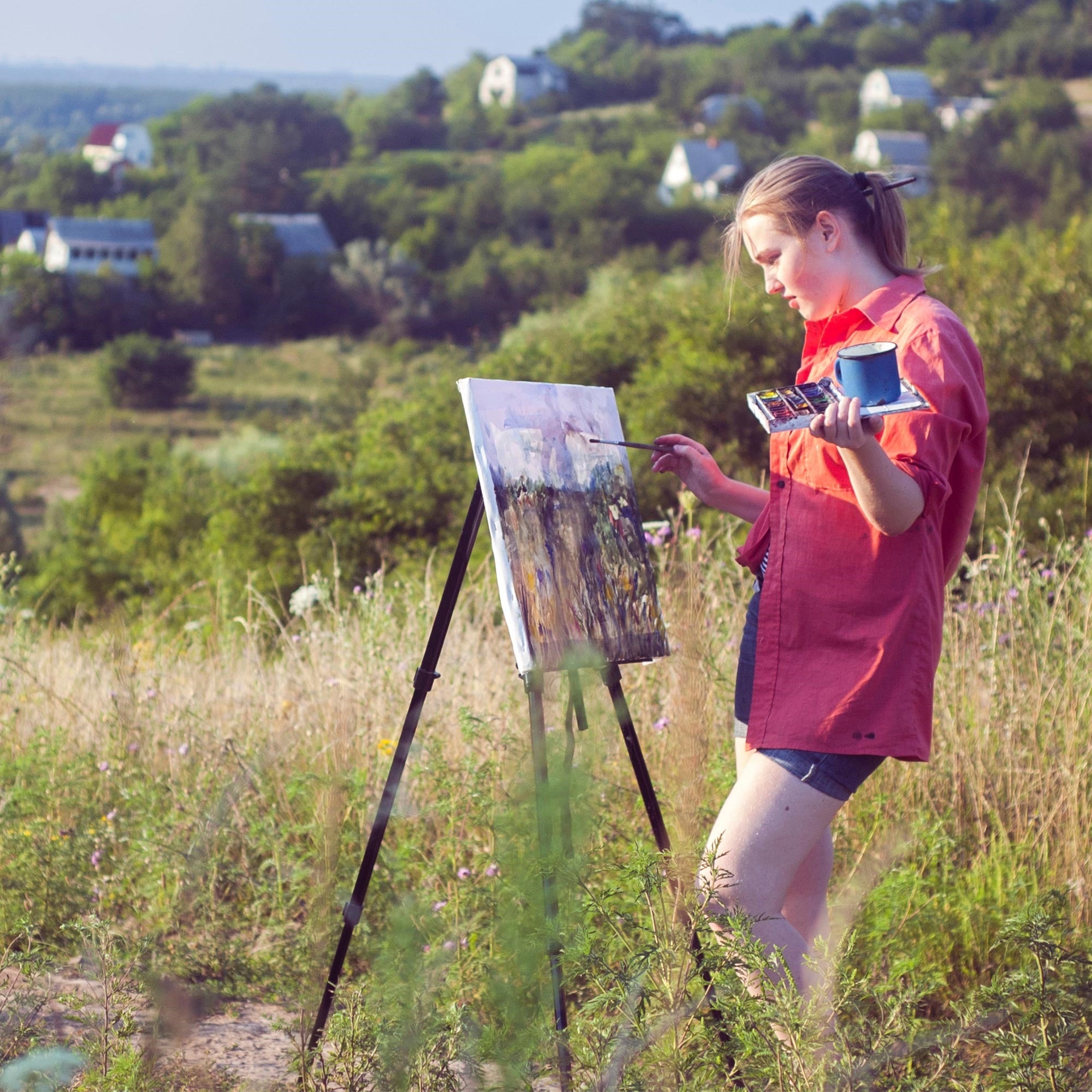 Frau malt an einer Staffelei in einer grünen Landschaft.