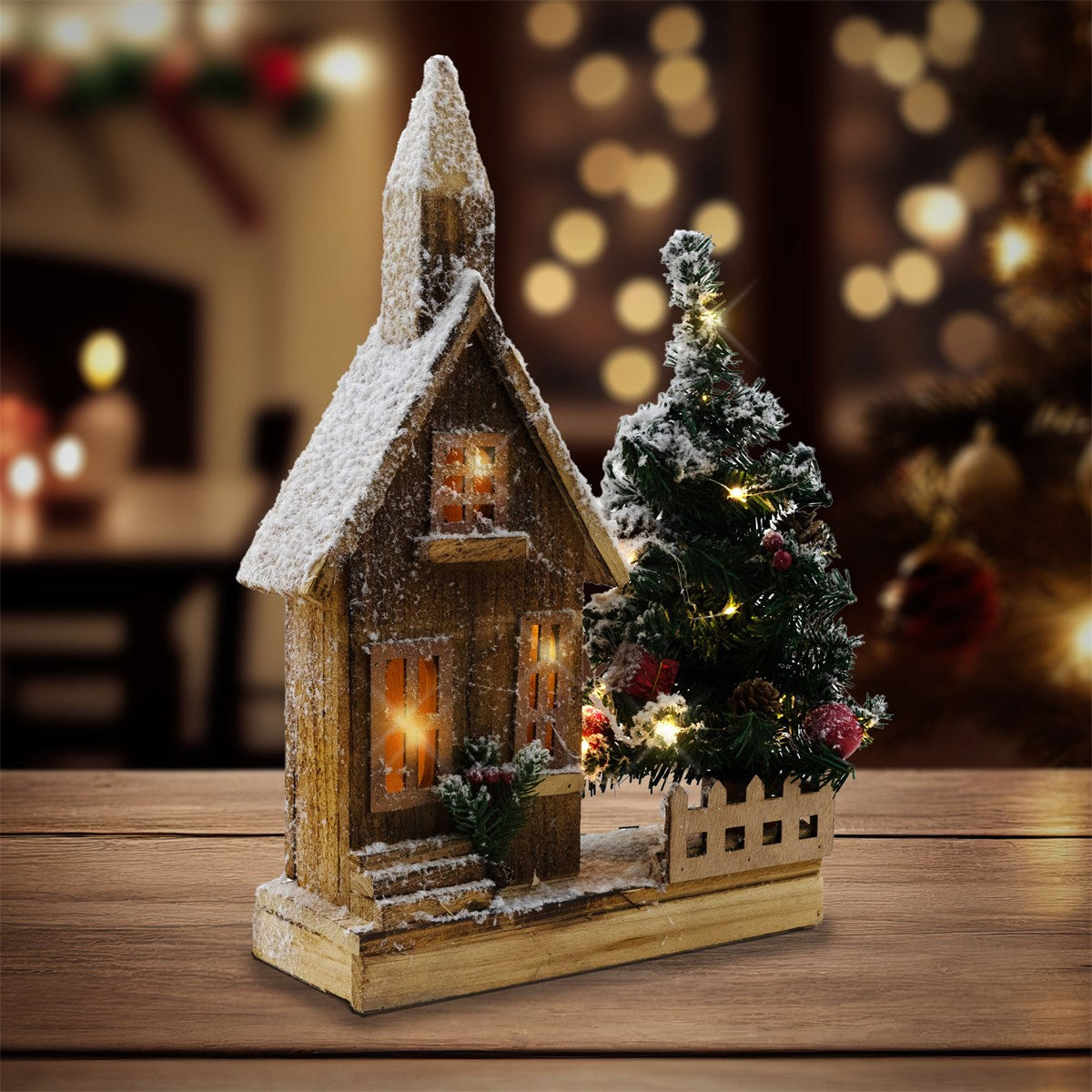 Dekoriertes Holzhaus mit schneebedecktem Dach und beleuchteten Fenstern neben einem geschmückten Tannenbaum.