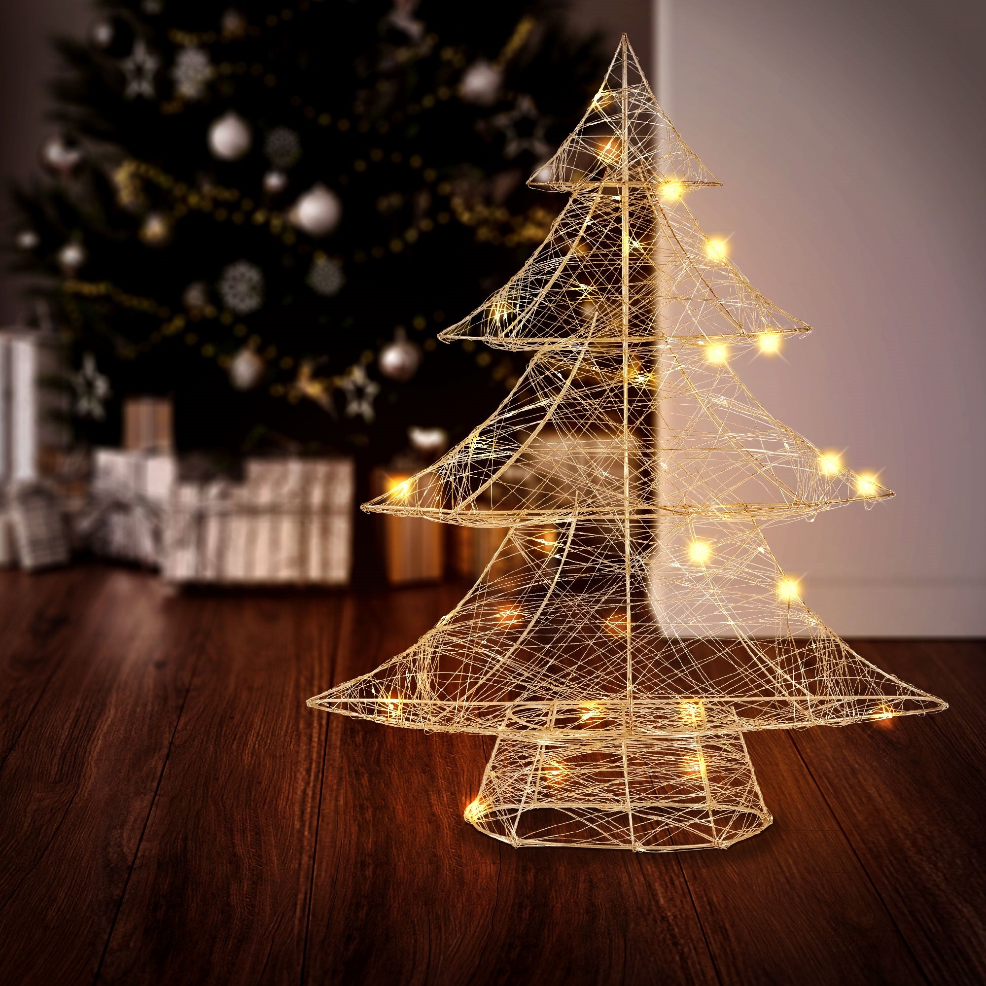 Ein beleuchteter Weihnachtsbaum aus Draht auf einem Holzboden vor einem Weihnachtsbaum.
