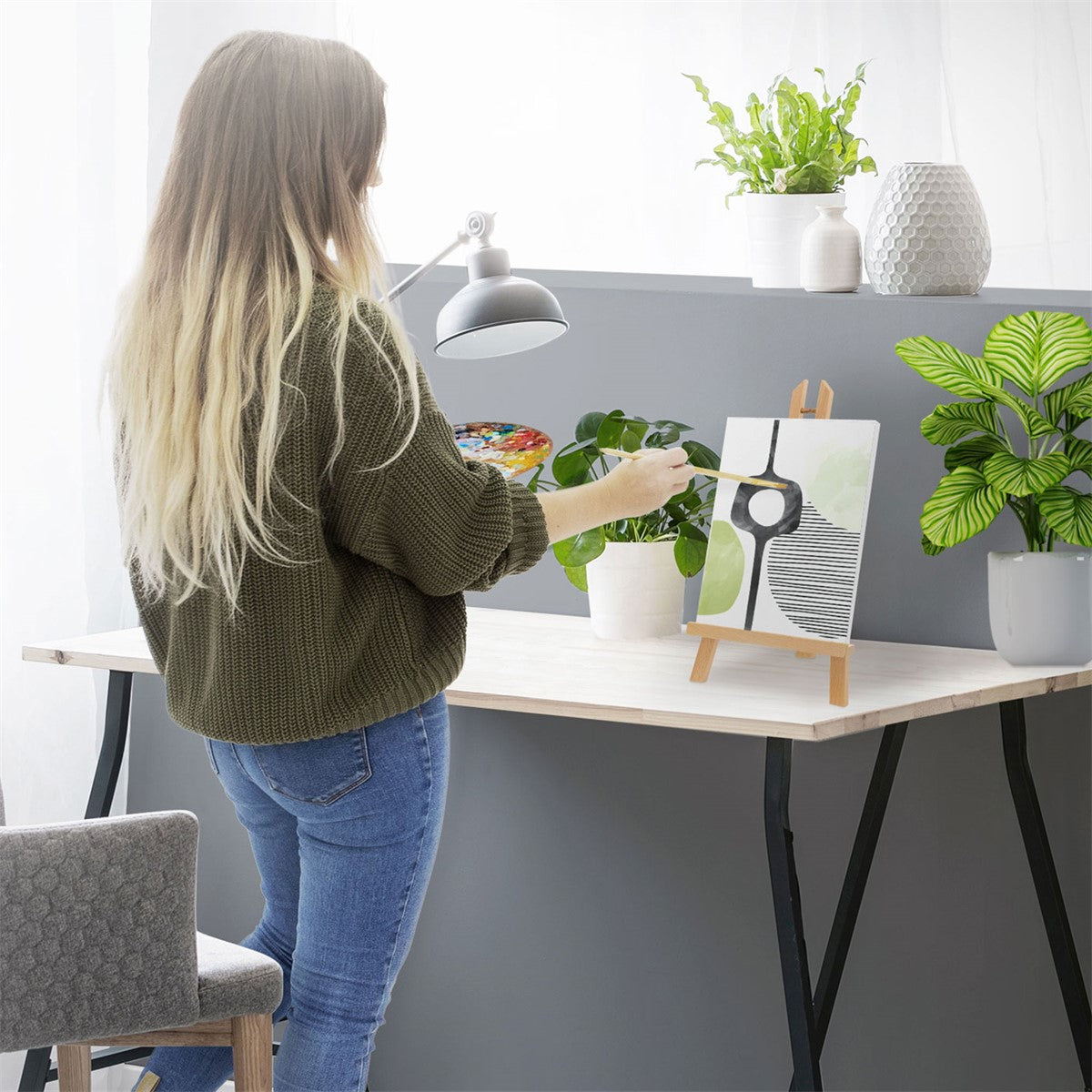 Frau malt an einem Tisch, Kunstwerk auf einem Ständer, Pflanzen im Hintergrund.