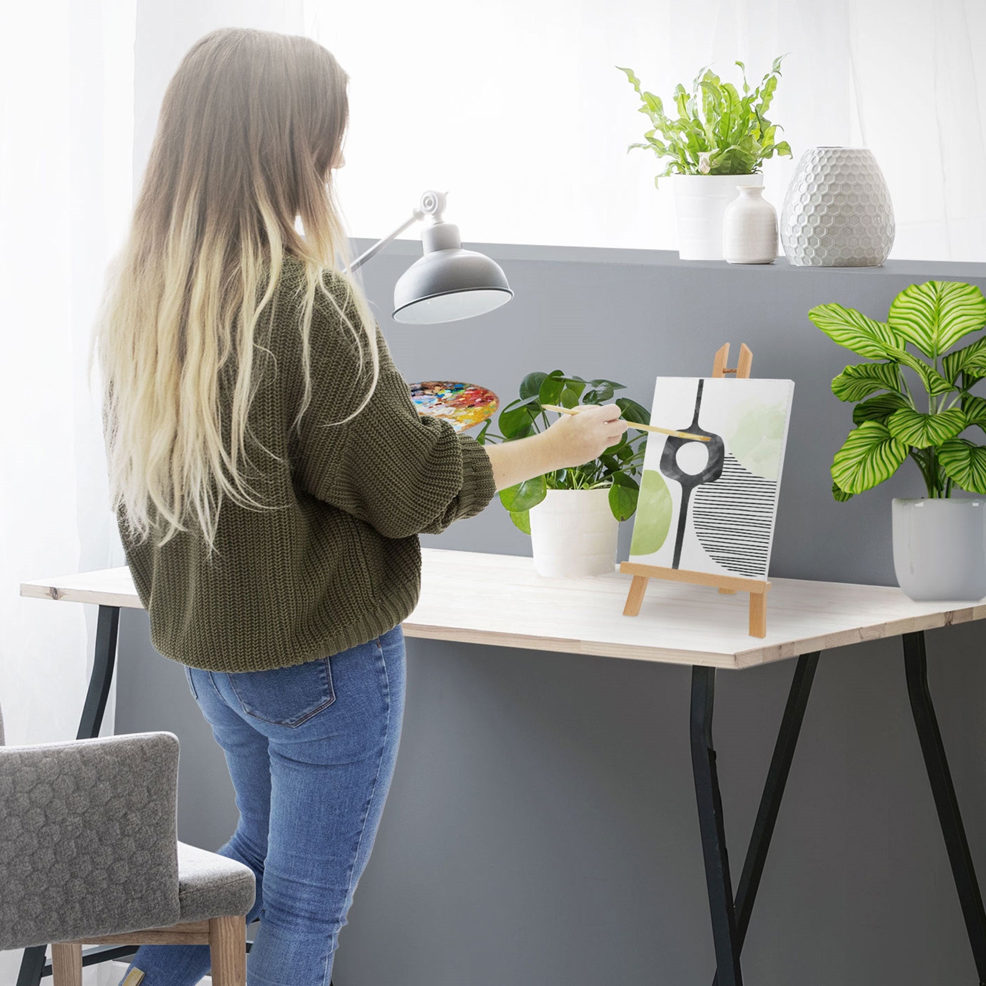 Frau malt an einem Tisch, Kunstwerk auf einem Ständer, Pflanzen im Hintergrund.
