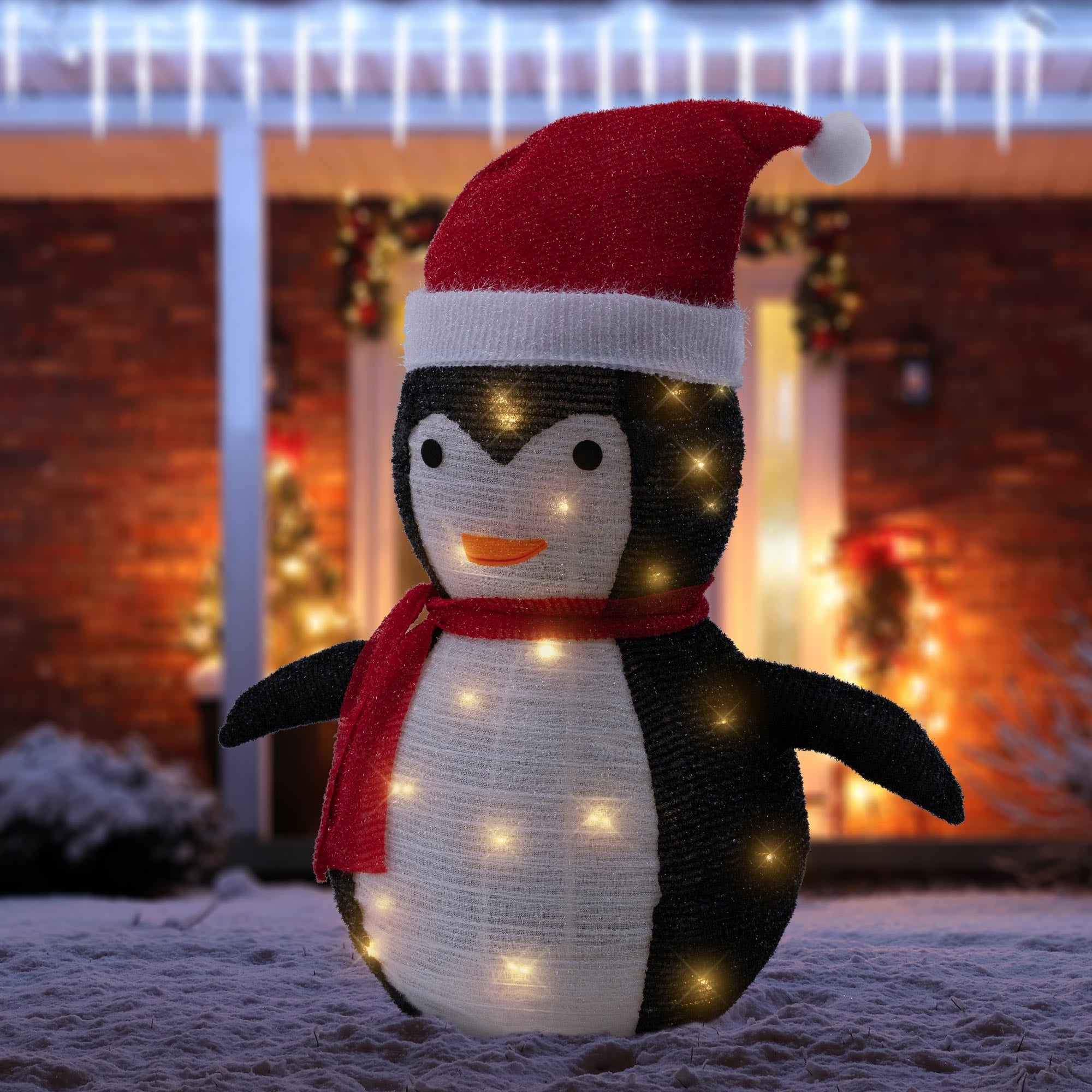 Ein leuchtender Pinguin mit rotem Hut und Schal vor einem festlich dekorierten Haus.