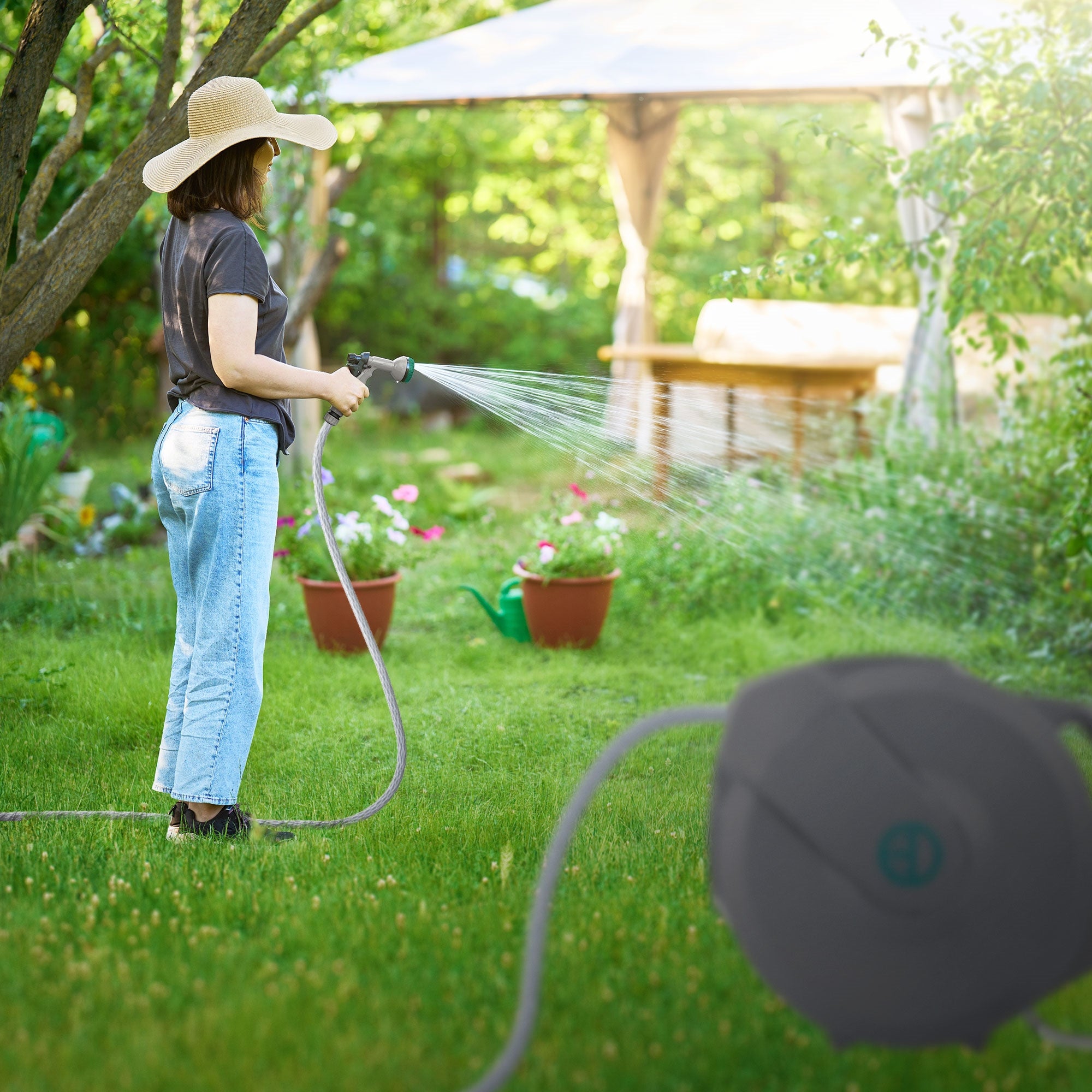 Frau mit Hut spritzt Wasser auf Pflanzen im Garten.