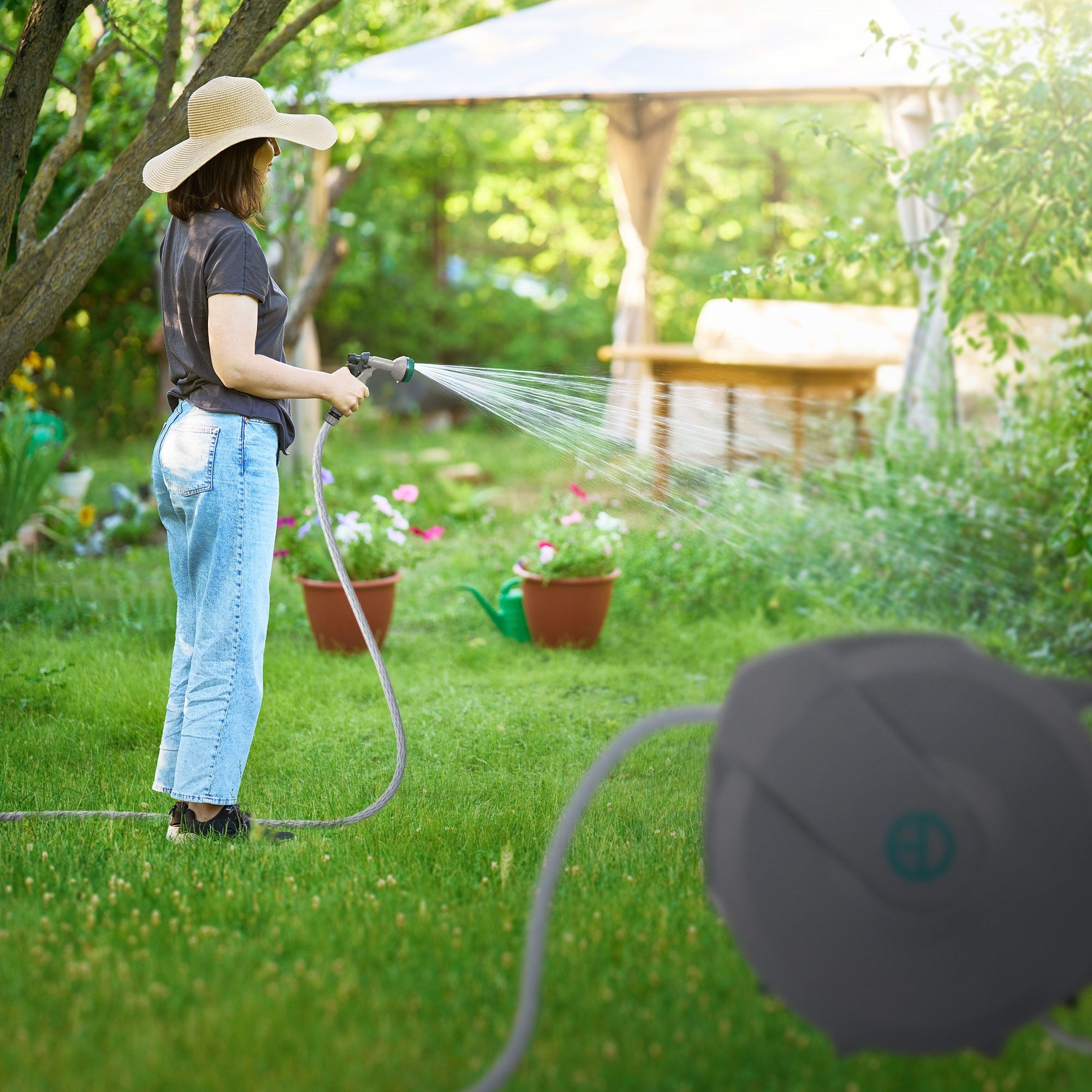 Frau im Garten spritzt Wasser mit einem Schlauch, im Hintergrund Pflanzen und ein Pavillon.