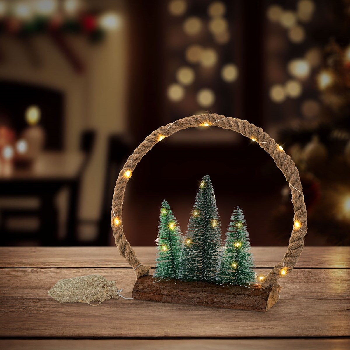 Beleuchteter Ring aus gedrehtem Seil auf einem Holzsockel, davor stehen drei kleine Tannenbäume.