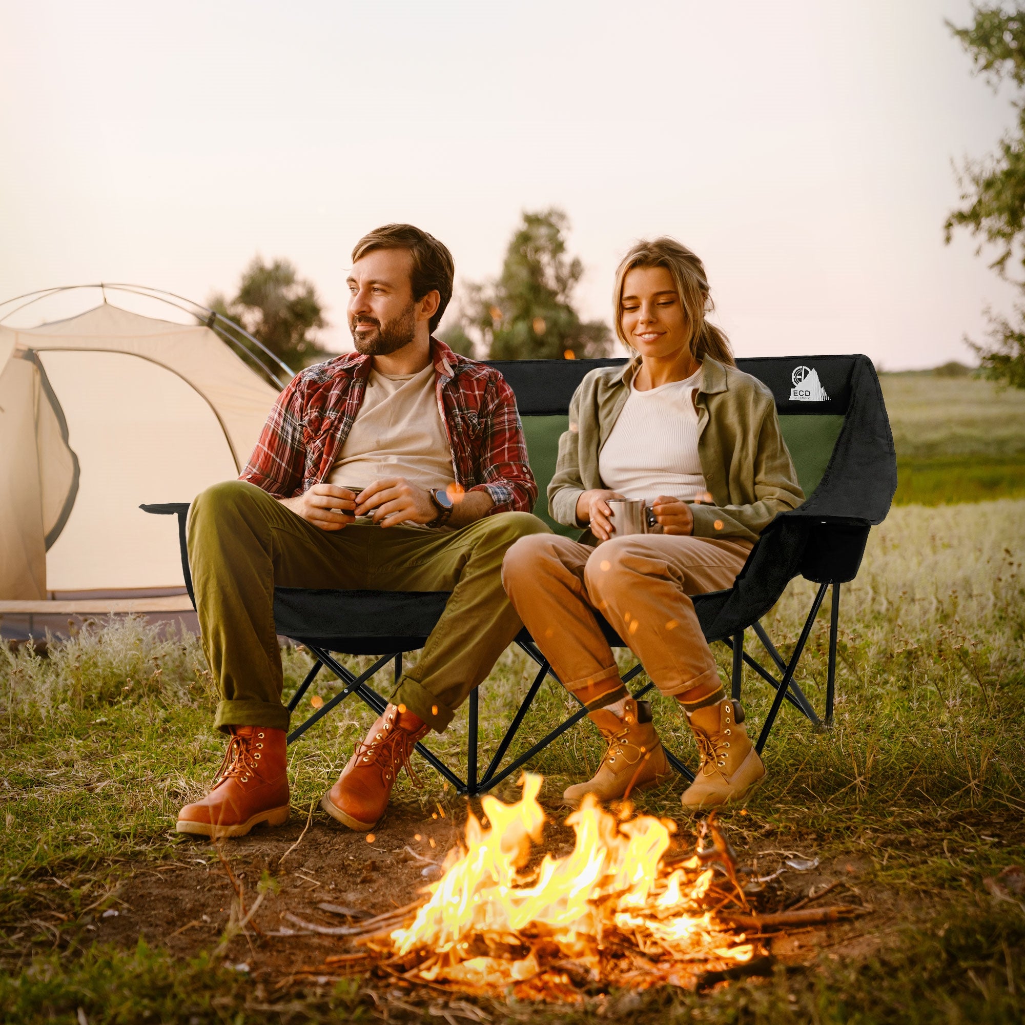 Paar sitzt am Lagerfeuer in Campingstühlen, Zelt im Hintergrund.