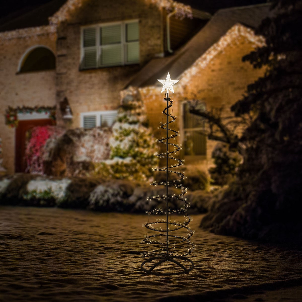 Beleuchteter Weihnachtsbaum im Freien vor einem Haus, mit Schnee im Hintergrund.