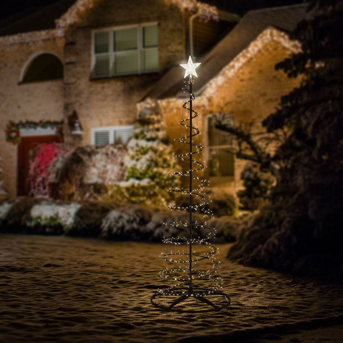 Beleuchteter Weihnachtsbaum im Freien vor einem Haus, mit Schnee im Hintergrund.