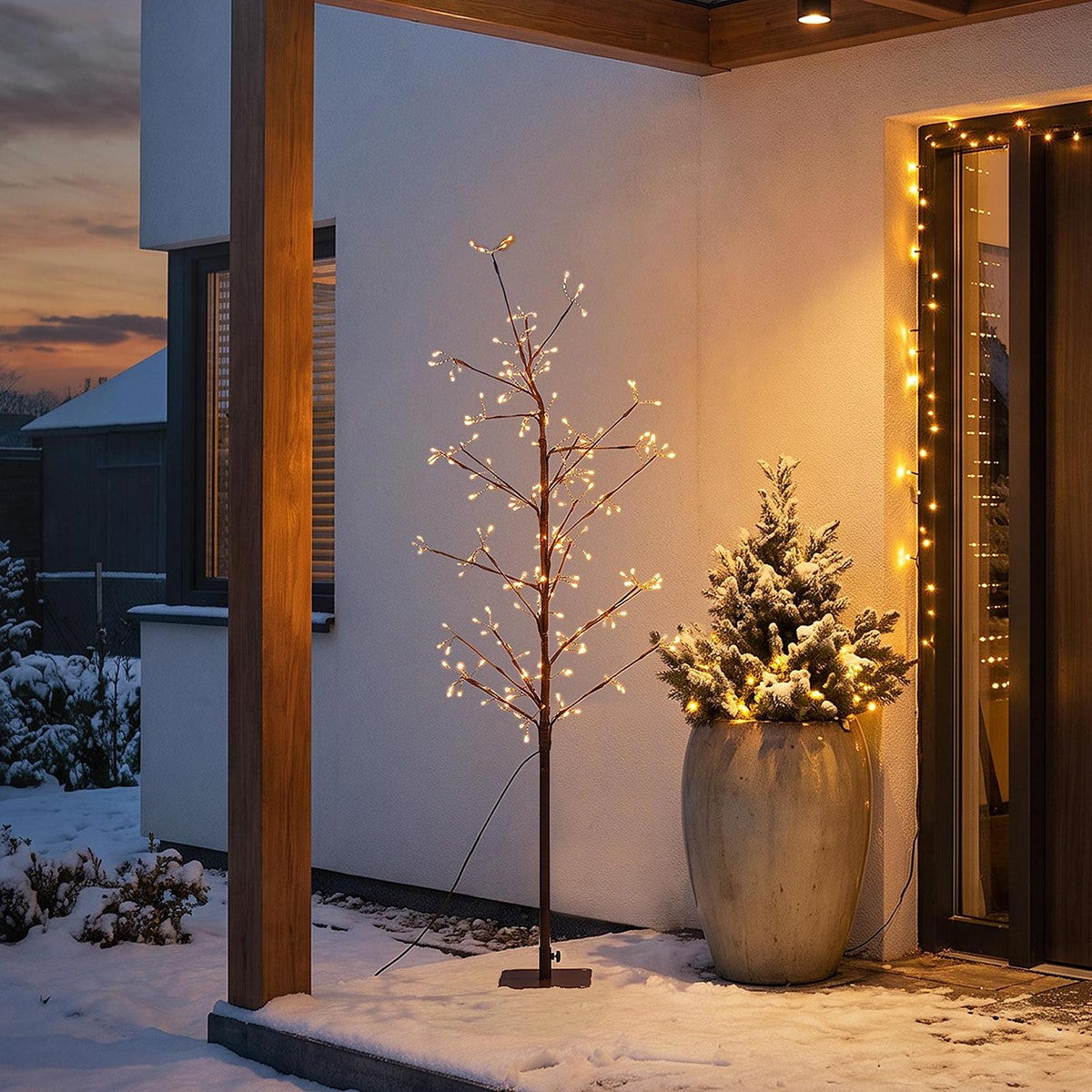 Beleuchteter künstlicher Baum neben einem bepflanzten Topf auf einer verschneiten Veranda bei Dämmerung.