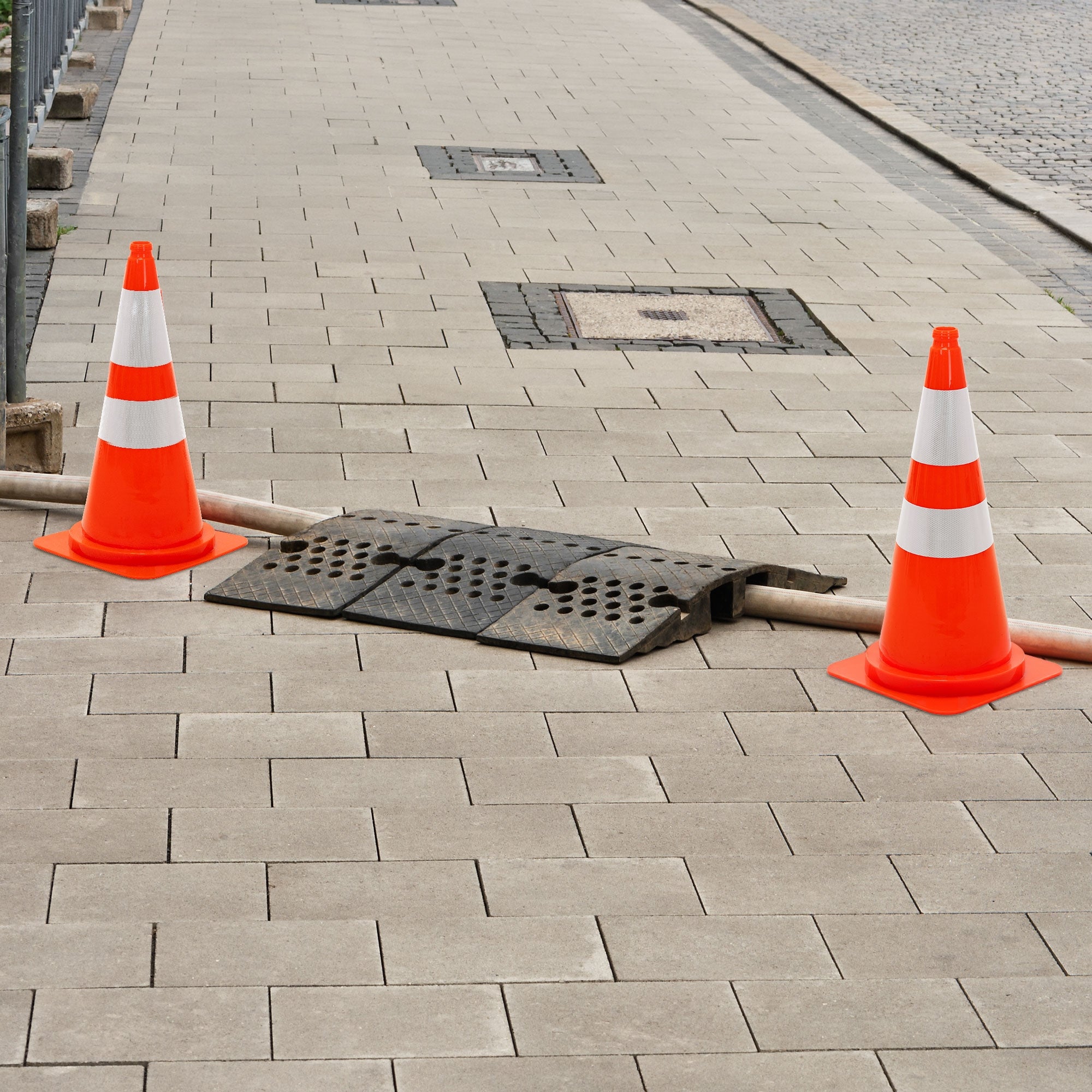 Zwei Verkehrsleitkegel neben einem Gitterrost auf einem Gehweg.