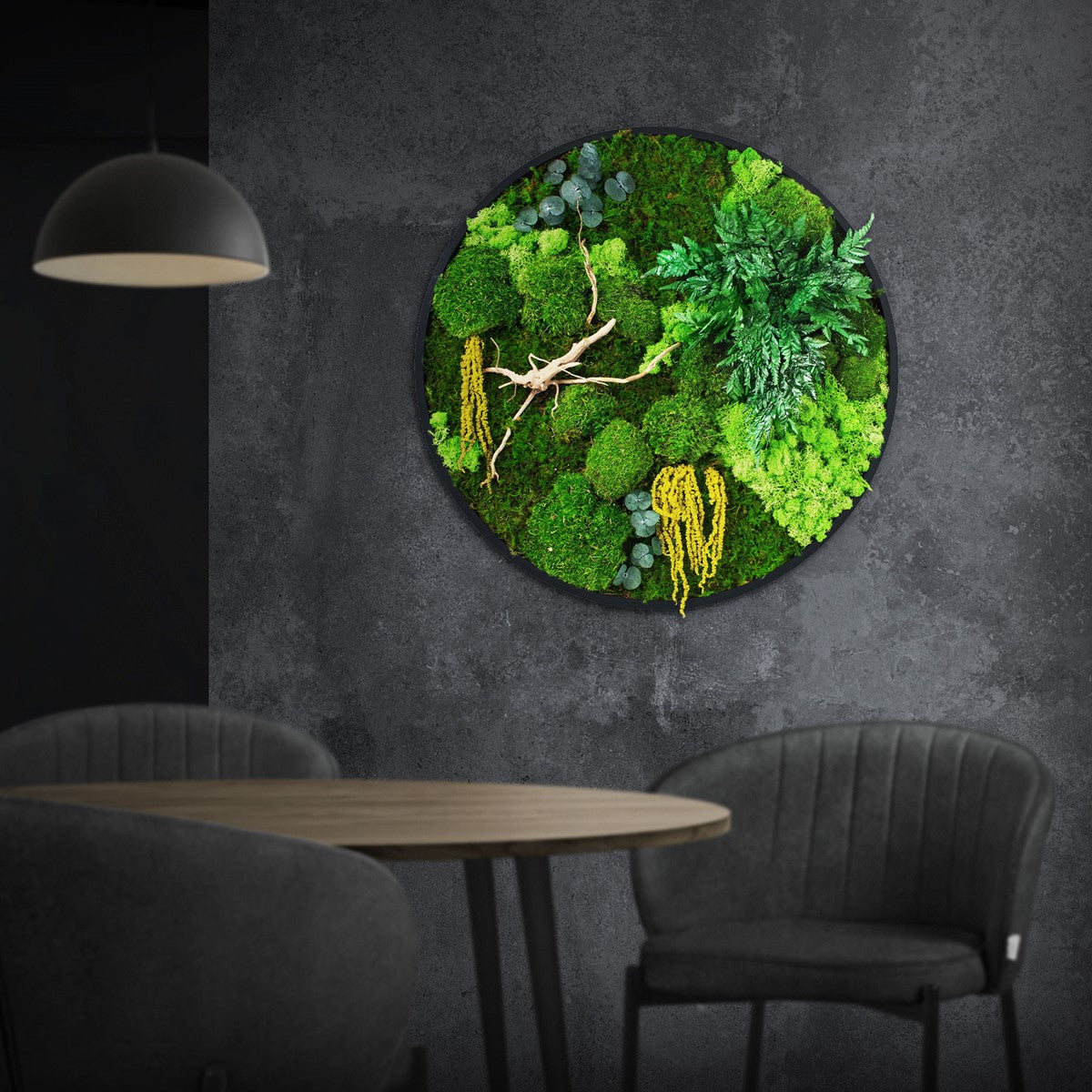 Rundes Moosbild mit verschiedenen grünen Moosen, Farn, Eukalyptus und einem Zweig hängt über Tisch und Stühlen an einer dunklen Wand.
