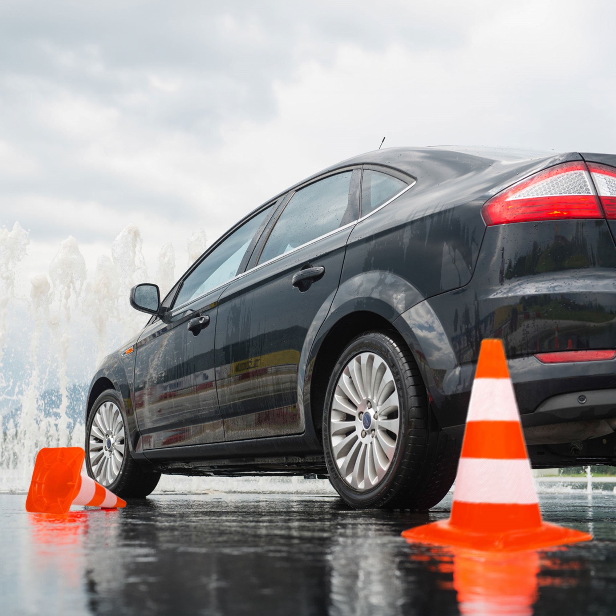 Schwarzes Auto neben orangefarbenen Pylonen auf nasser Straße mit Spritzwasser im Hintergrund.