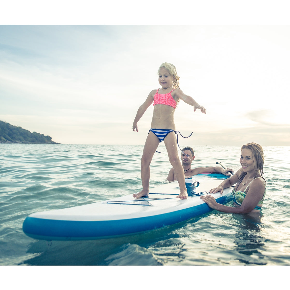 Ein Kind steht auf einem Surfbrett im Wasser, umgeben von zwei Erwachsenen.