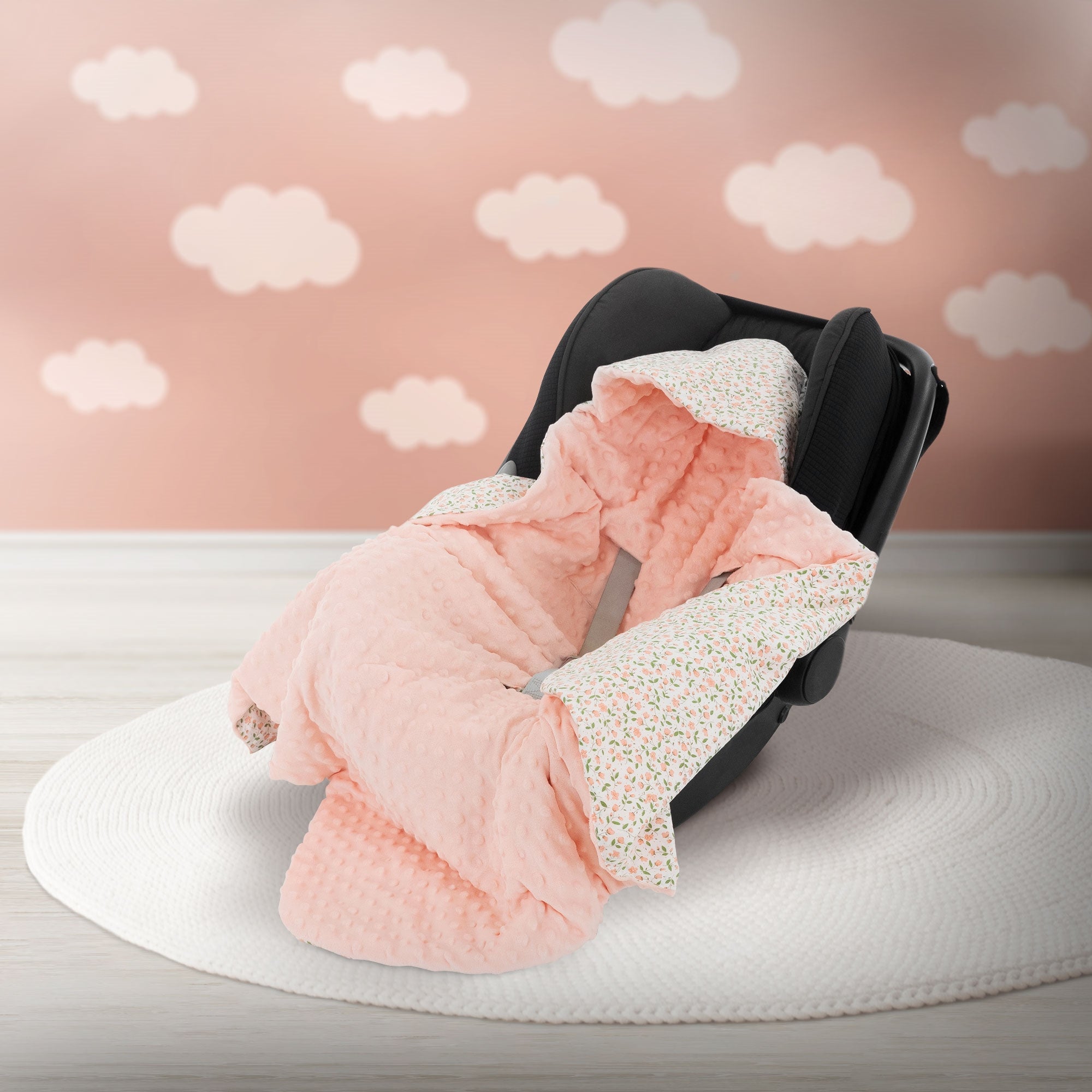 Un siège auto noir avec une couverture rose sur un tapis rond devant un mur rose avec des nuages.