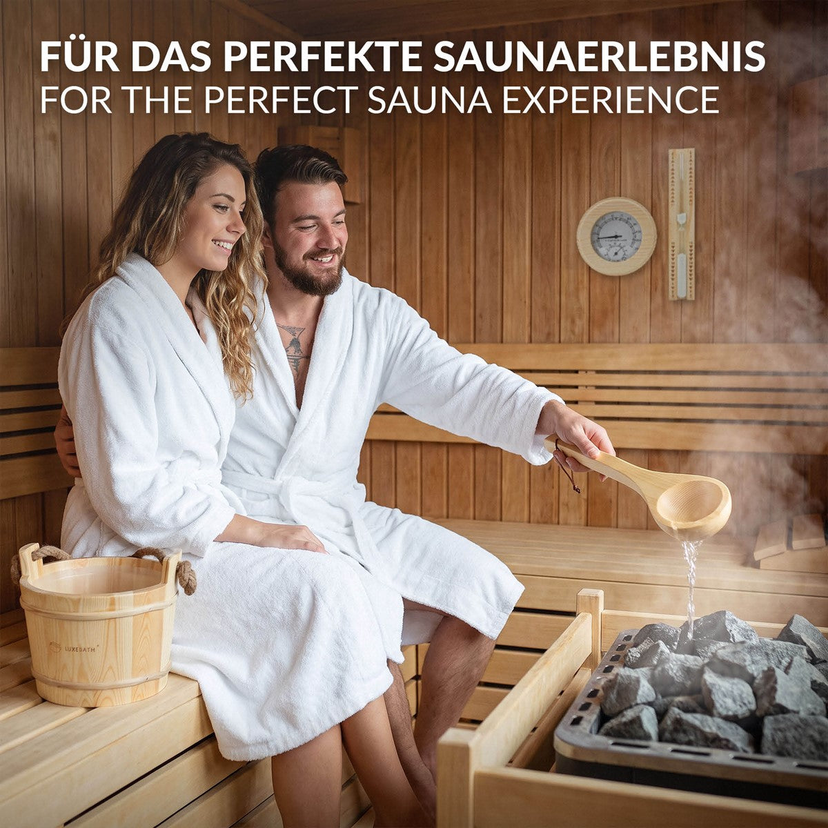 Paar in Bademänteln sitzt in einer Sauna, Holzofen im Hintergrund, Wasserschaufel in der Hand.