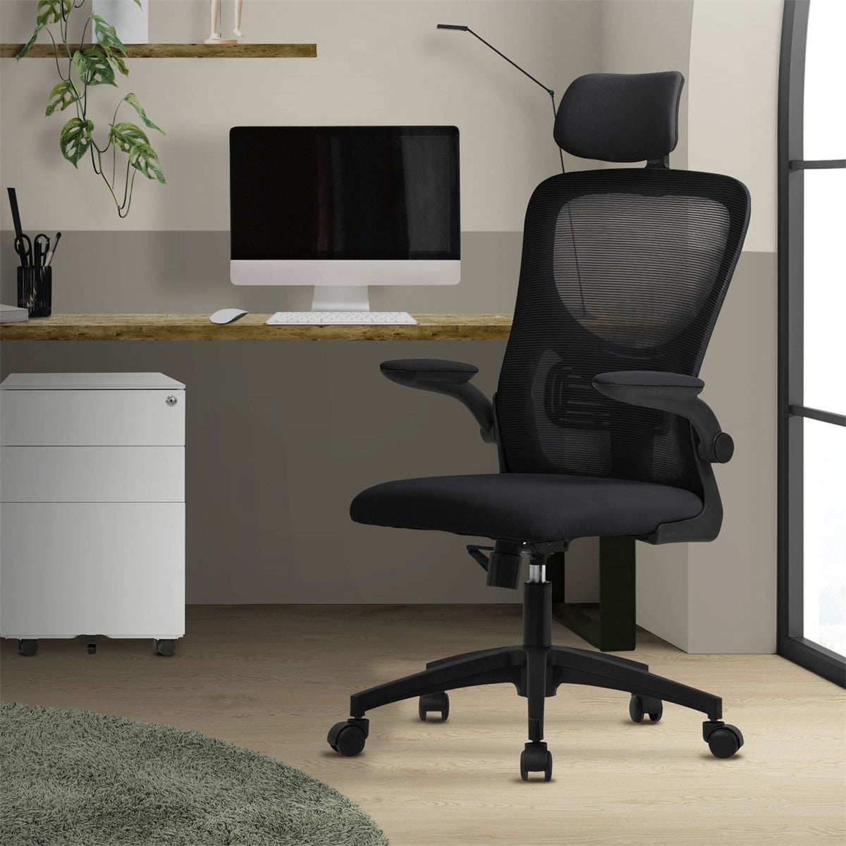 A black office chair in front of a desk, with a computer and plants in the background.