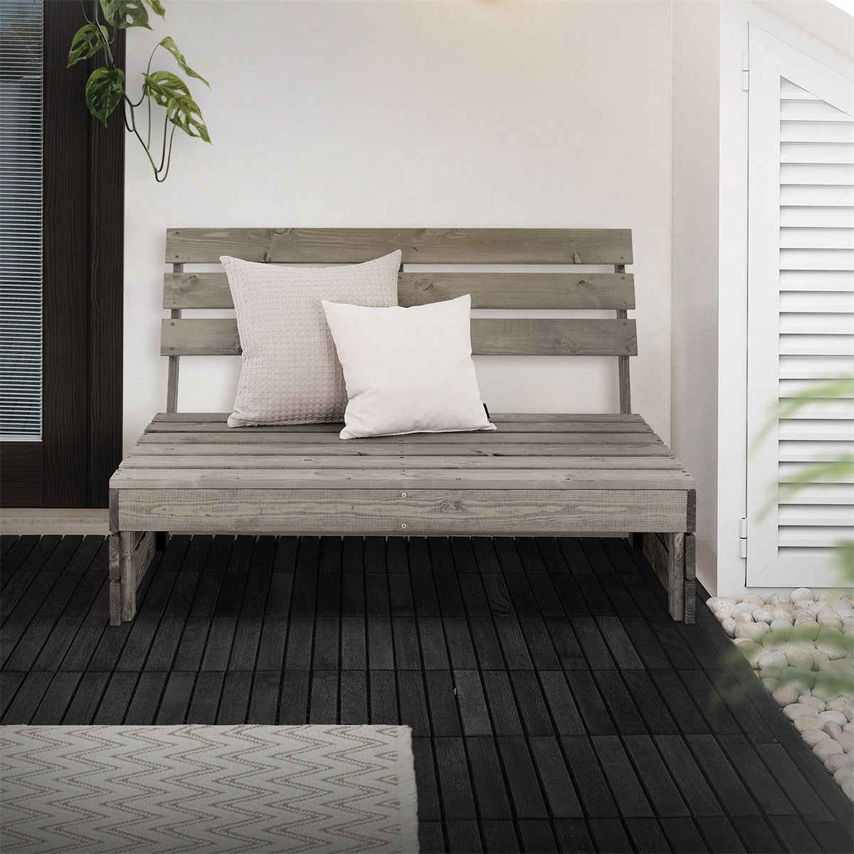 Gray wooden bench with two cushions, in front of a wall and plants in the background.