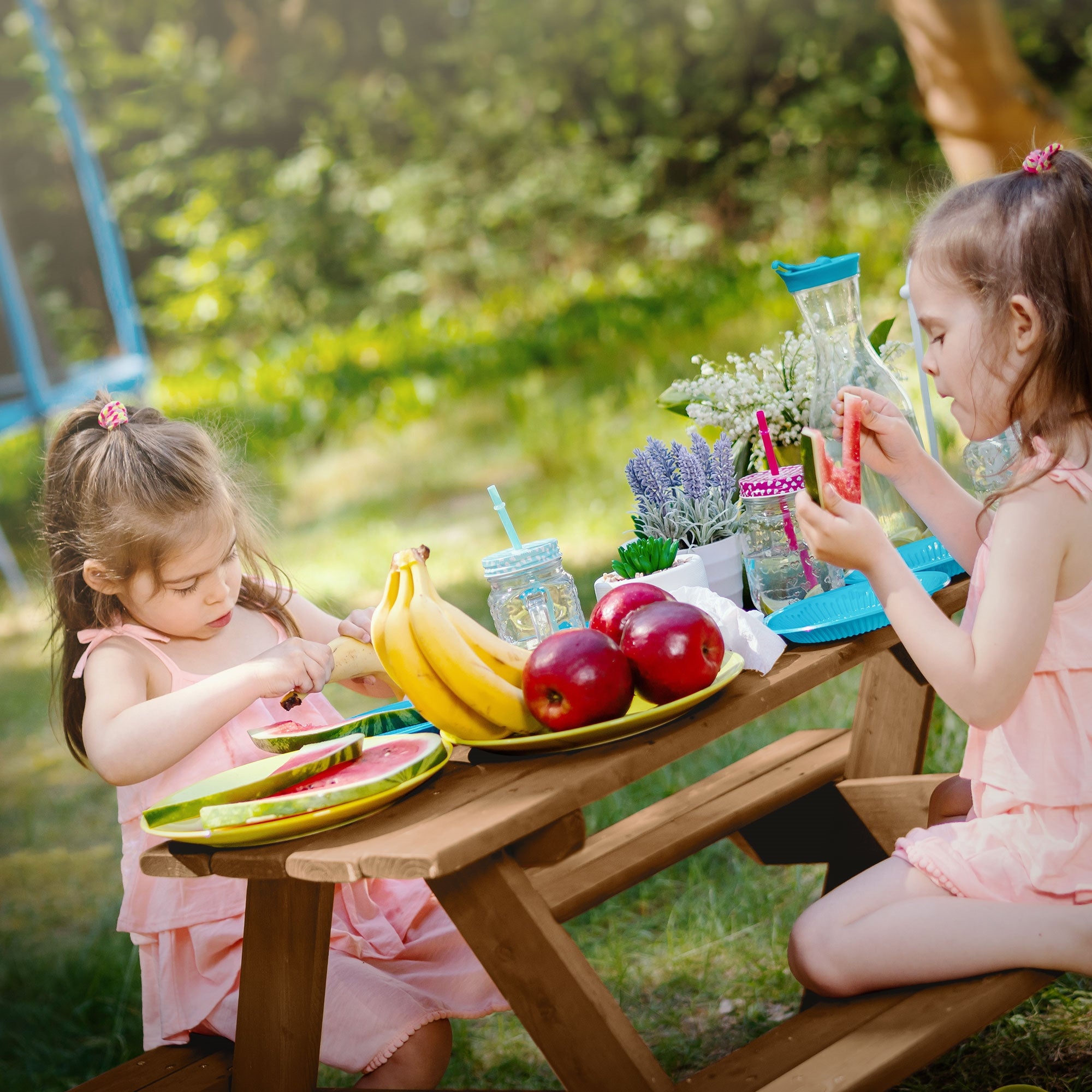 Zwei Kinder sitzen an einem Tisch und essen Obst, umgeben von Spielzeug und Pflanzen.