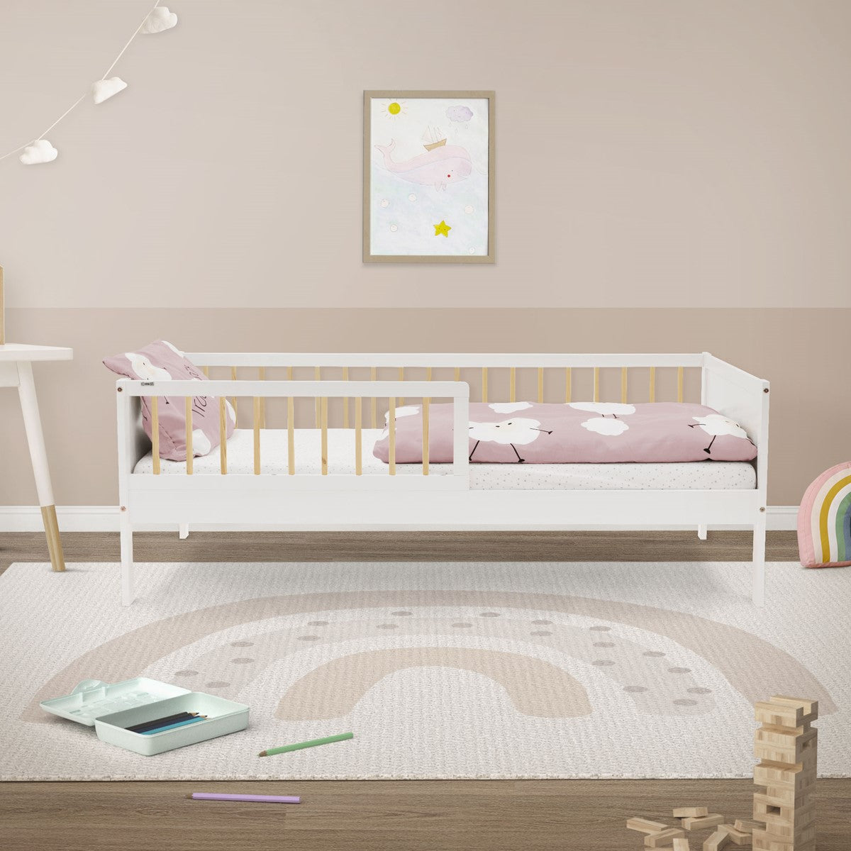Weißes Kinderbett mit rosa Bettwäsche in einem hellen Zimmer; auf dem Boden liegen ein Regenbogenteppich und Holzklötze.