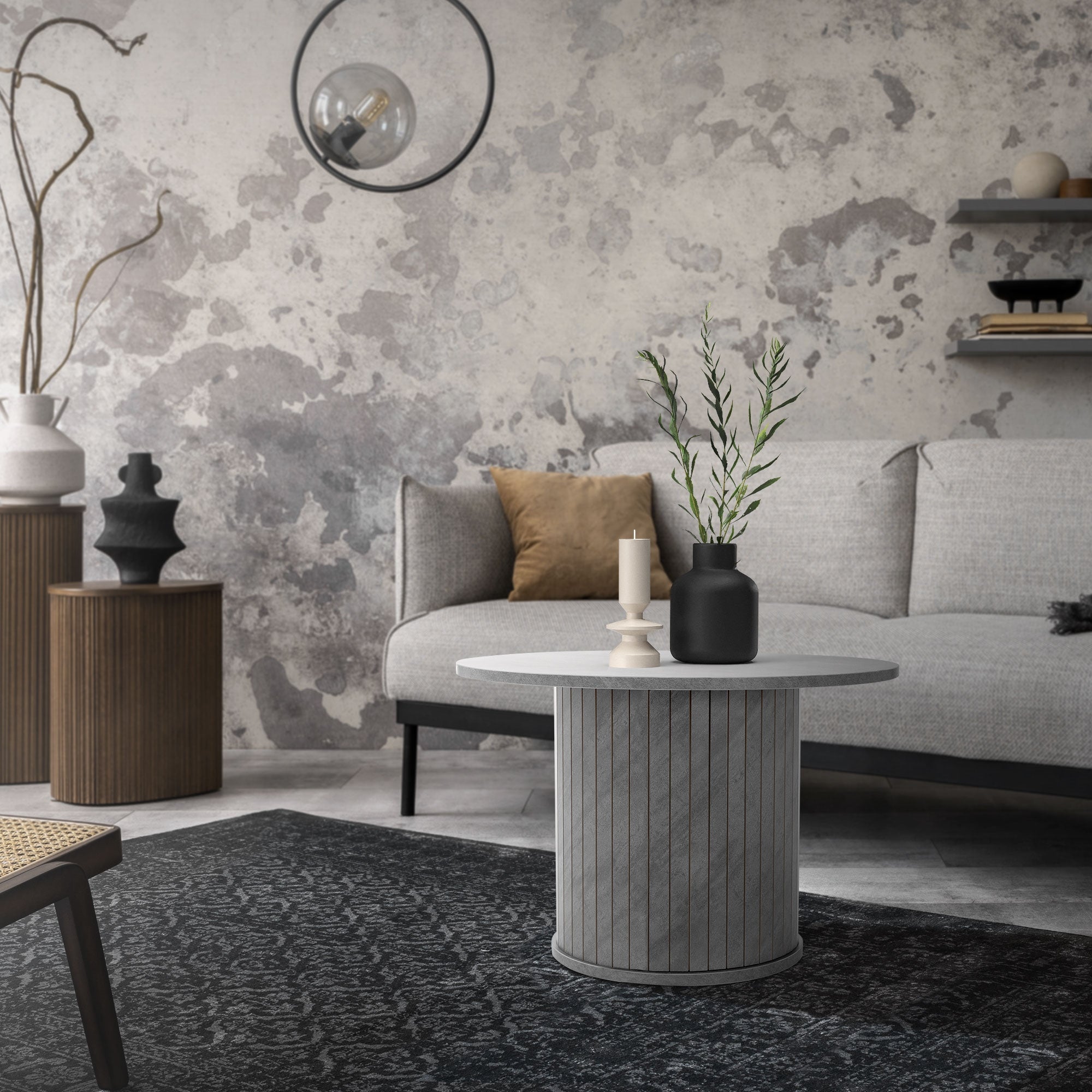 Stue med grå sofa, rundt bord og dekorative planter på et tæppe.