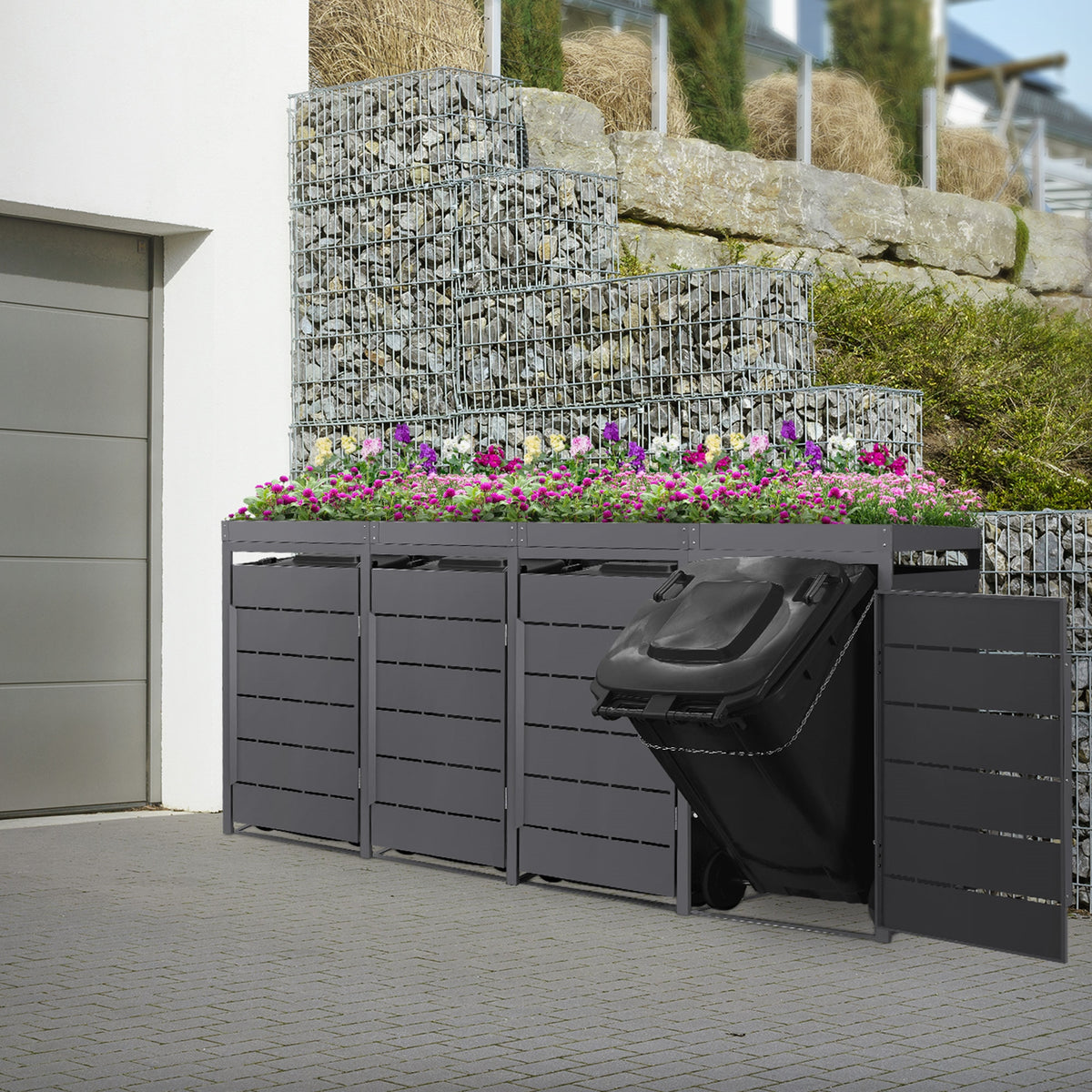 Mülltonnenbox mit Blumen, an einer Steinterrasse.