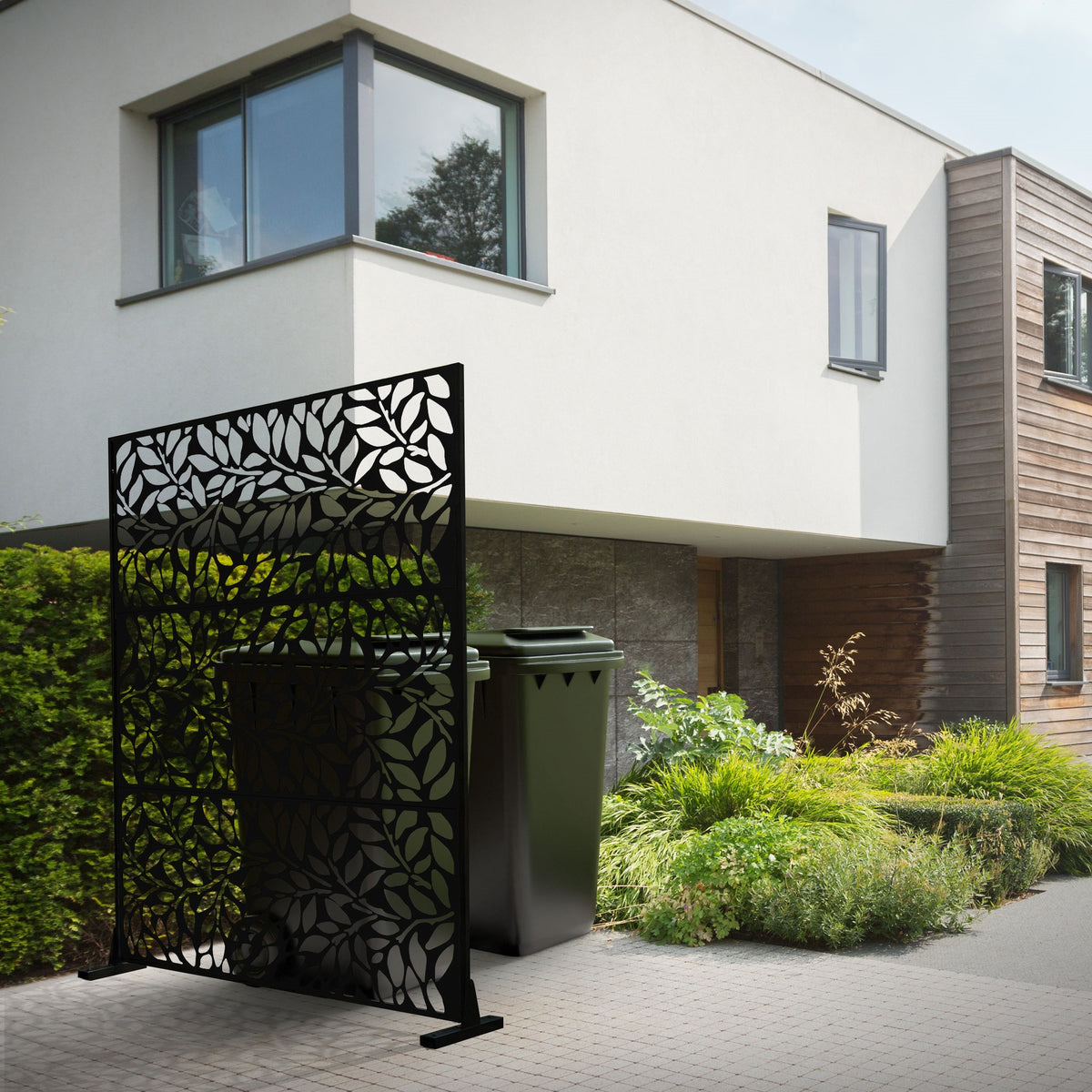 Sort skærmvæg med bladmønster foran en moderne husfacade og affaldsbeholdere.