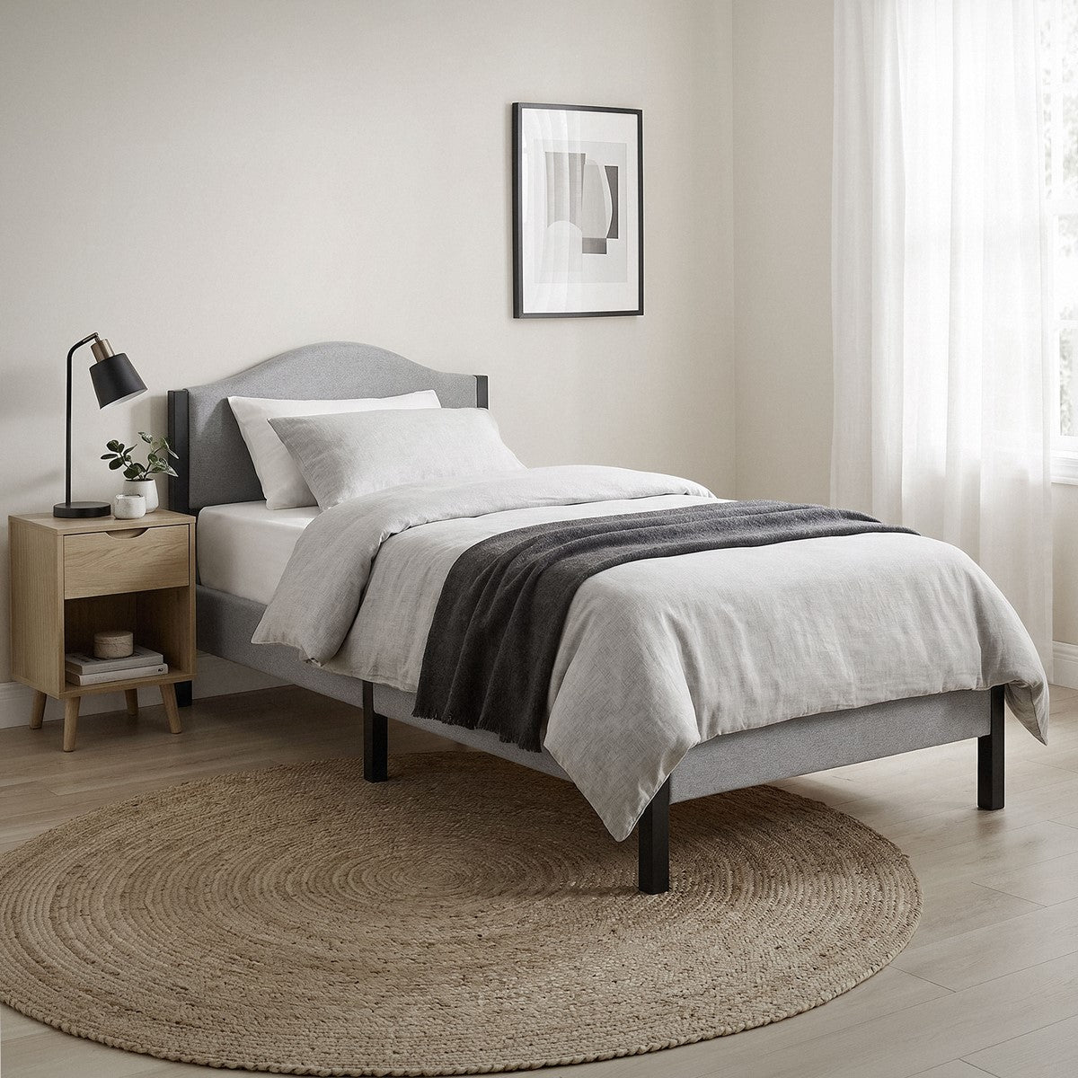 Letto grigio con biancheria da letto bianca e grigio scuro, comodino e tappeto rotondo intrecciato.