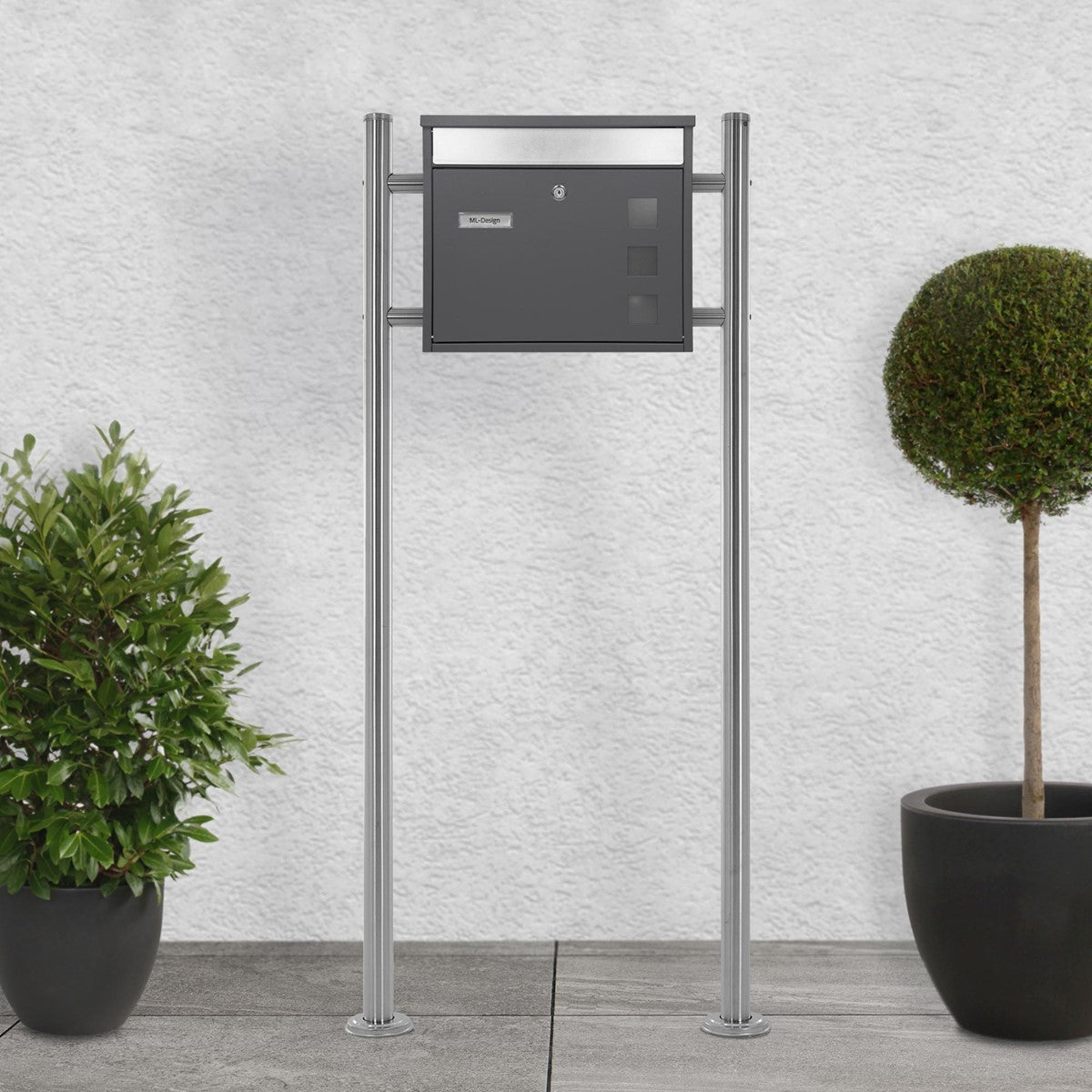 Mailbox on a metal stand, gray, in front of a wall with plants.