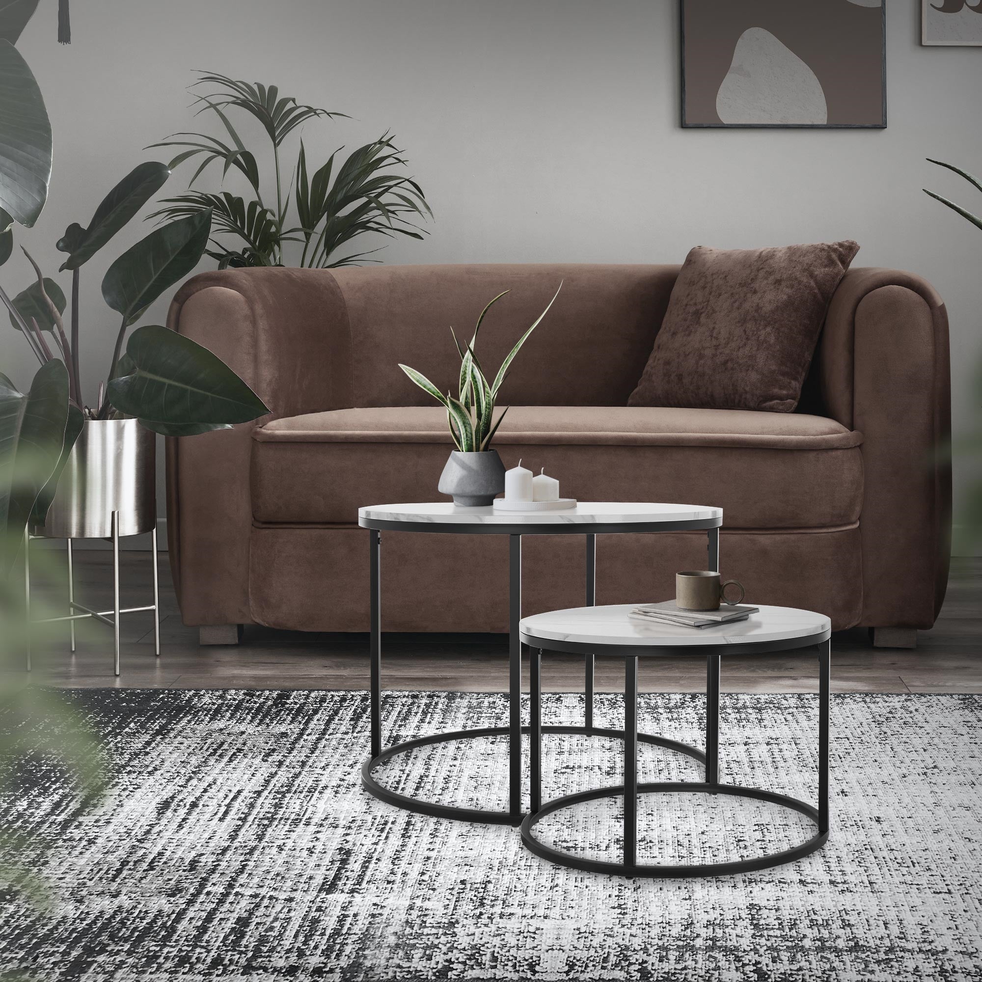 Wohnzimmer mit braunem Sofa, zwei Tischen und Pflanzen im Hintergrund.