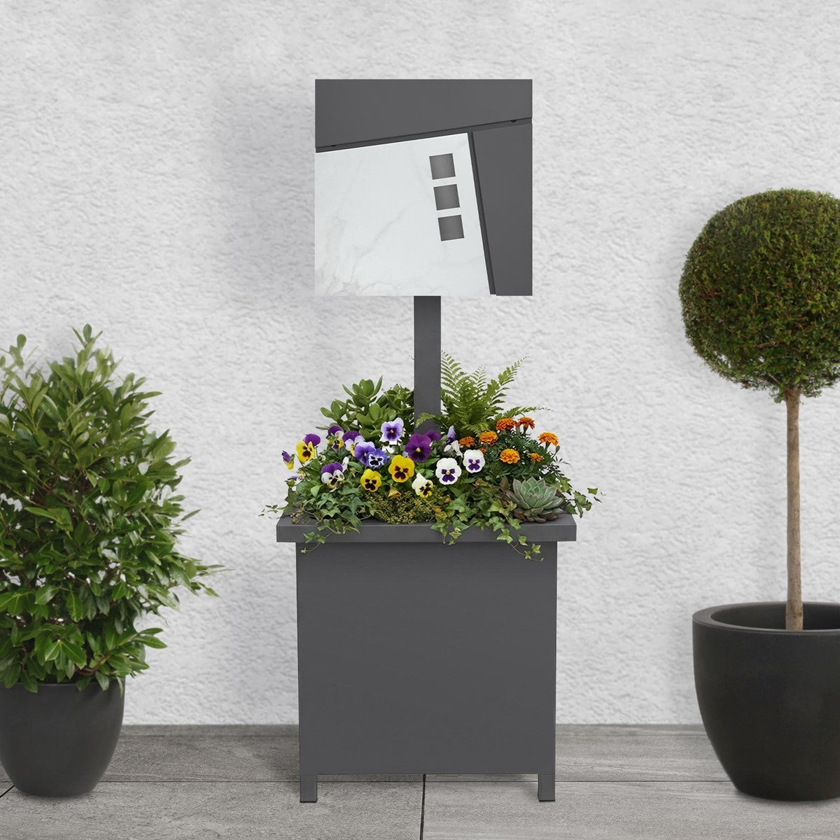 Briefkasten auf einem Pflanzgefäß mit bunten Blumen, vor einer grauen Wand.