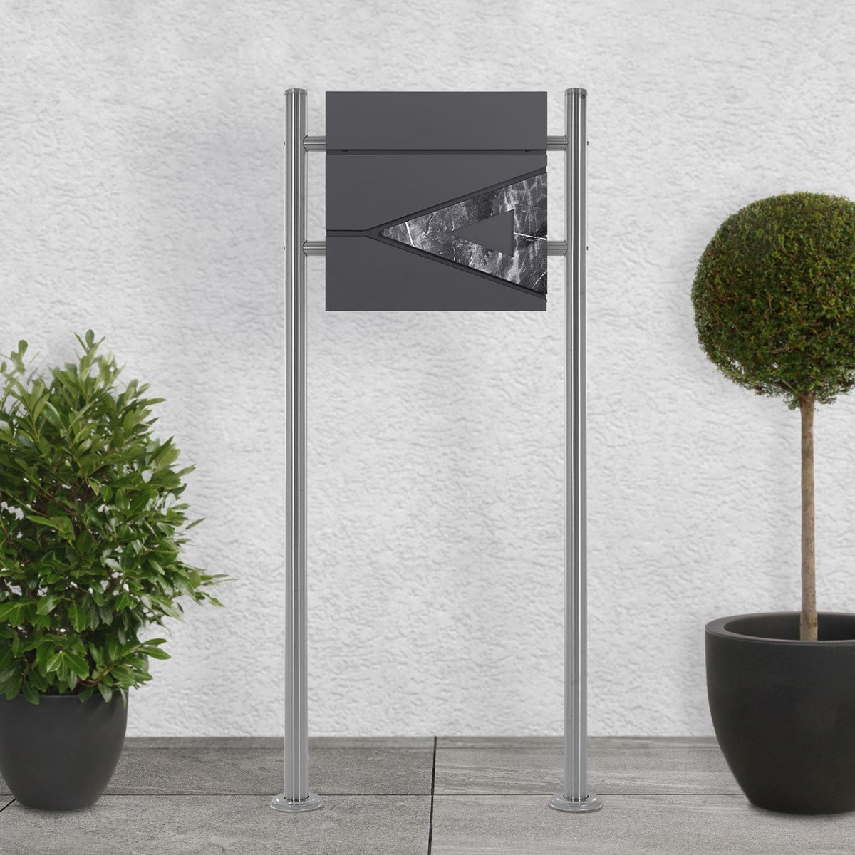Mailbox with a gray front and triangular design on a sidewalk with plants.