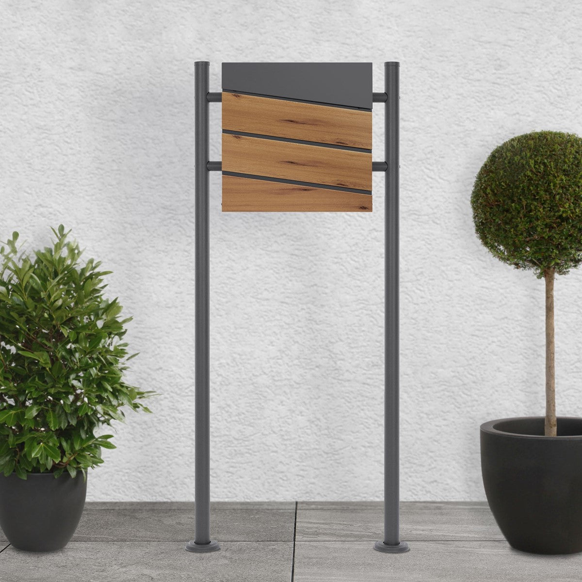 Mailbox with a wooden front and a grey frame, surrounded by two plants on a walkway.
